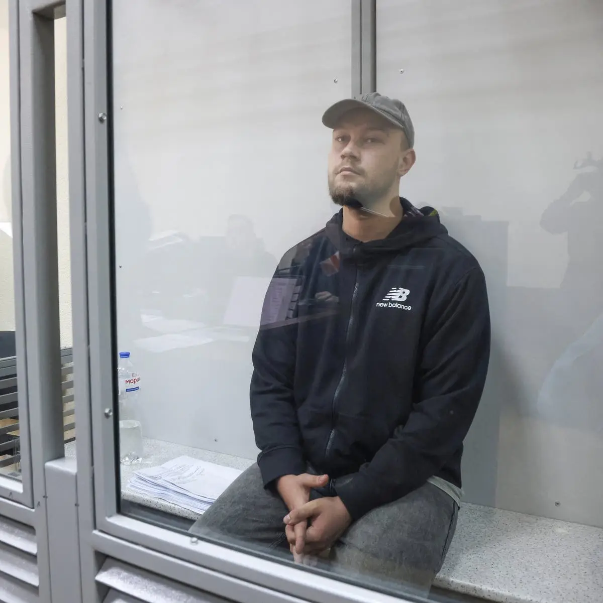 A man in a black New Balance hoodie and grey cap sits calmly inside a glass-enclosed booth, looking directly at the camera…