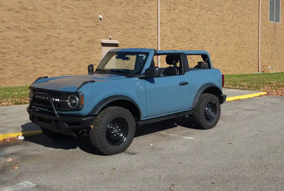 A light blue Ford Bronco with black trim, large off-road tires, and a black front bumper guard is parked in an asphalt lot…