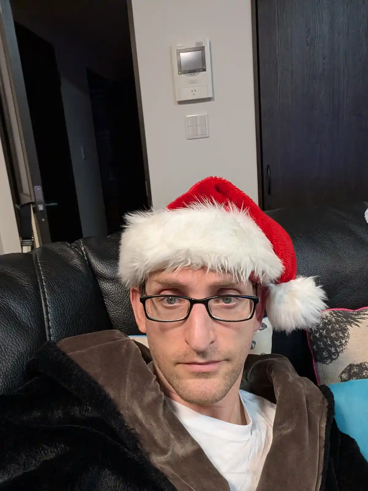 A man with glasses and a brown robe sits on a black leather couch wearing a red and white Santa hat. He has a tired, deadpan…