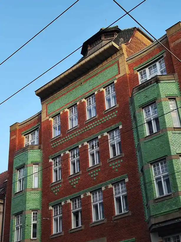 A low-angle photo shows the upper floors of a red brick building with green tiled accents and white windows against a clear…
