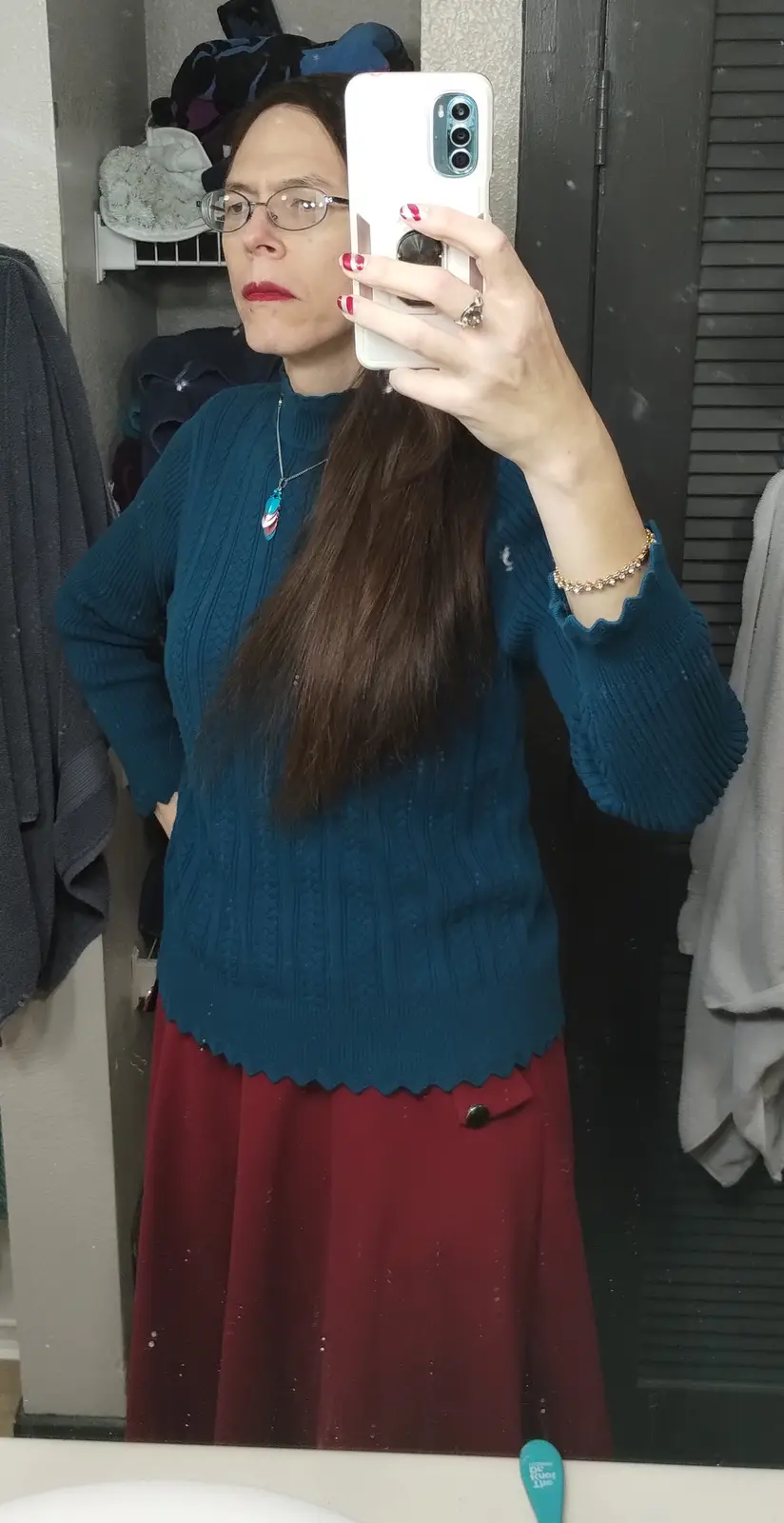 A woman with glasses and red lipstick takes a mirror selfie in a teal cable-knit sweater and red skirt, holding a white…