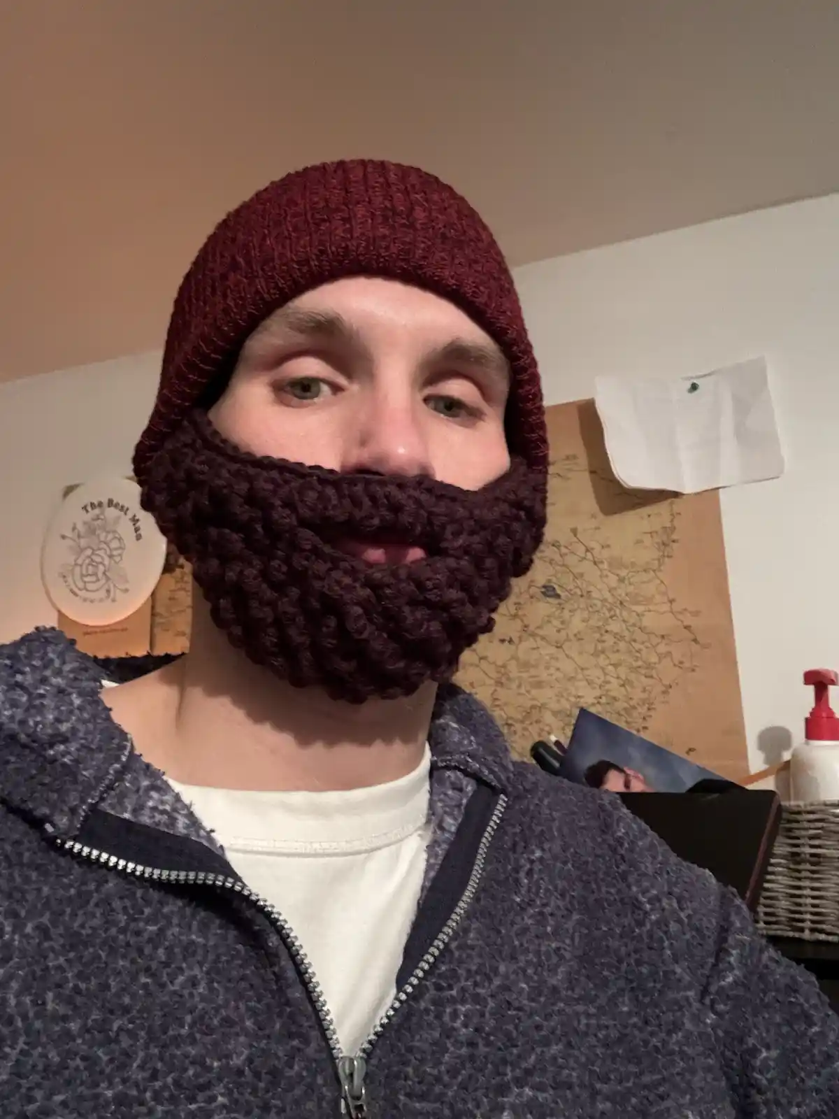 A man wearing a maroon knit beanie and a matching crocheted beard covering his lower face, looking directly at the camera…