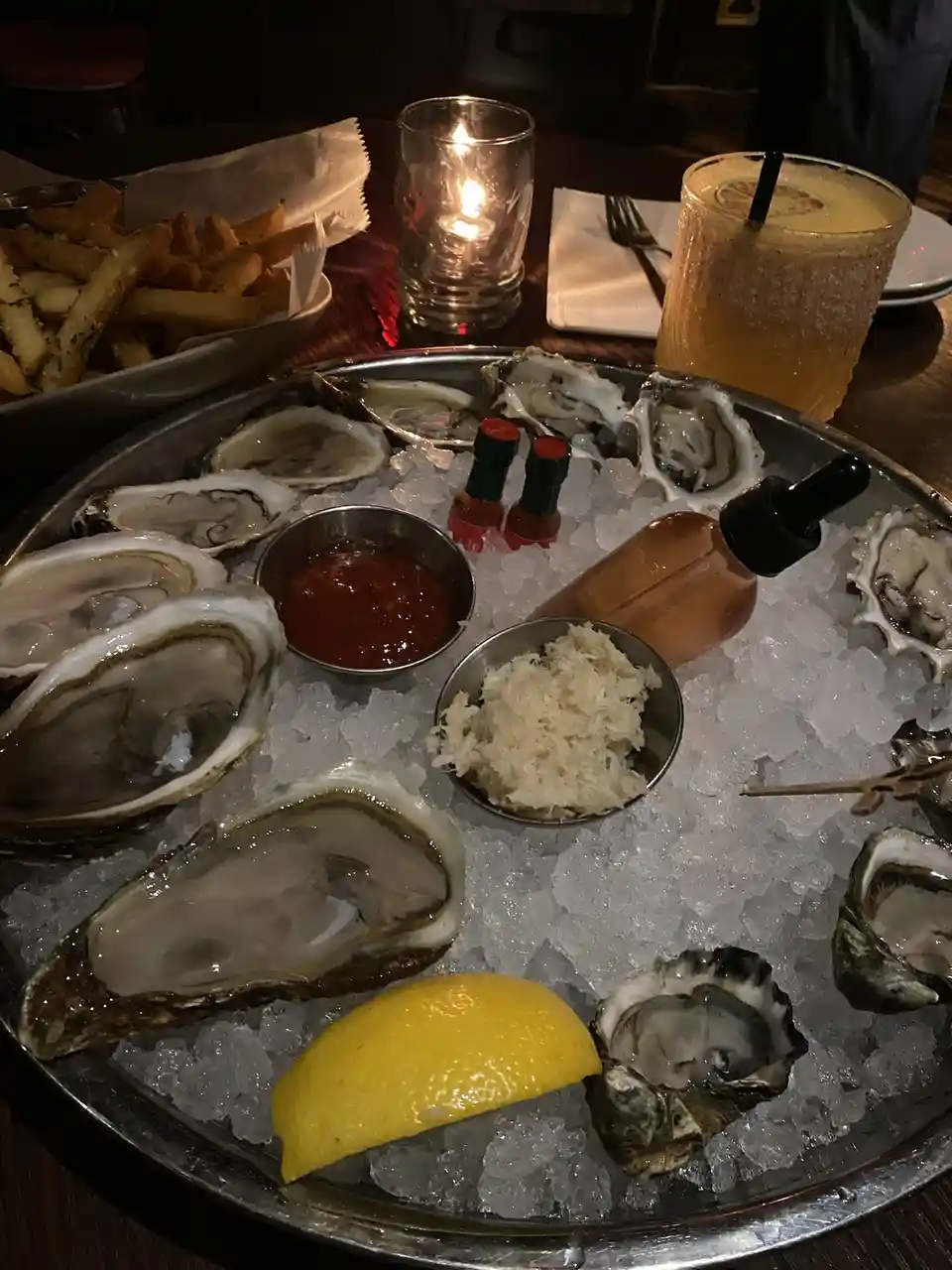 A metal tray filled with raw oysters on the half shell resting on crushed ice, accompanied by a lemon wedge, two small bowls…