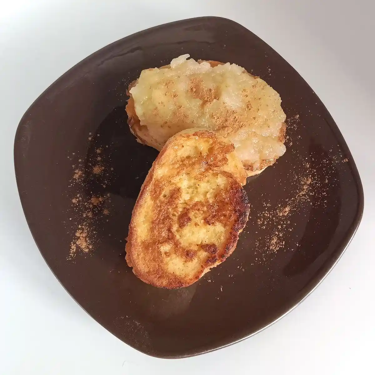 A dark brown, squarish plate holds two slices of toasted bread. One slice is topped with a spread that looks like mashed…
