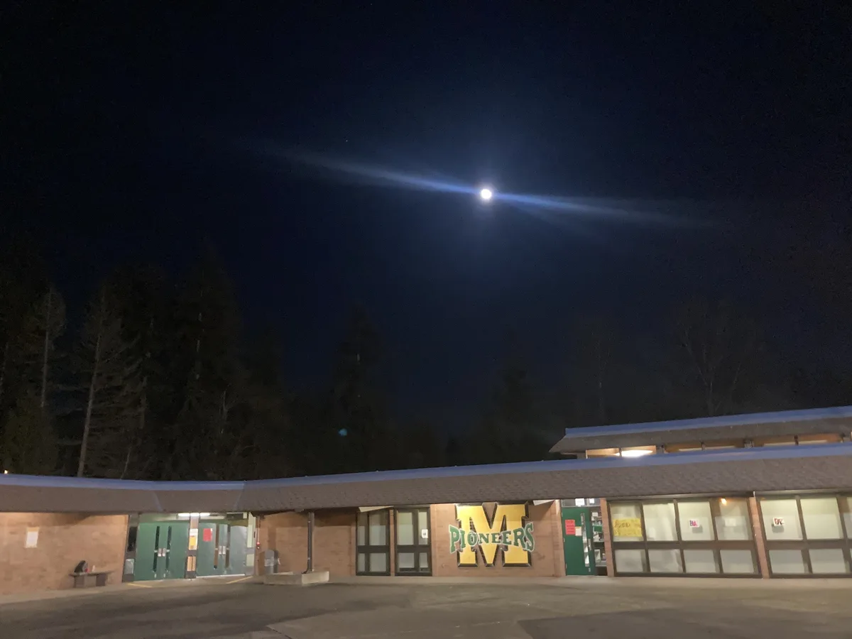 A nighttime photo of a school building with a large 'M PIONEERS' sign on its brick facade, under a dark sky where a bright…