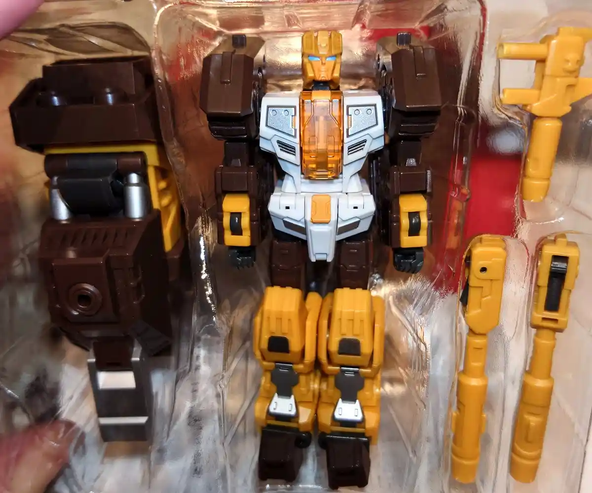 A close-up photo of a Transformers toy in its clear plastic packaging, showing the robot form with brown and yellow parts…