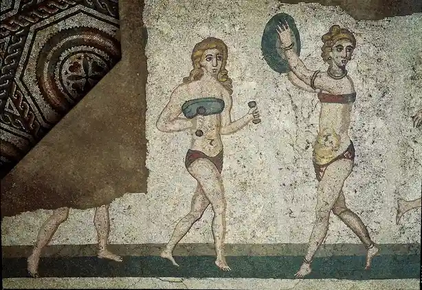 An ancient mosaic depicts two women in bikinis holding dumbbells and a shield, with a large section of the mosaic peeled…