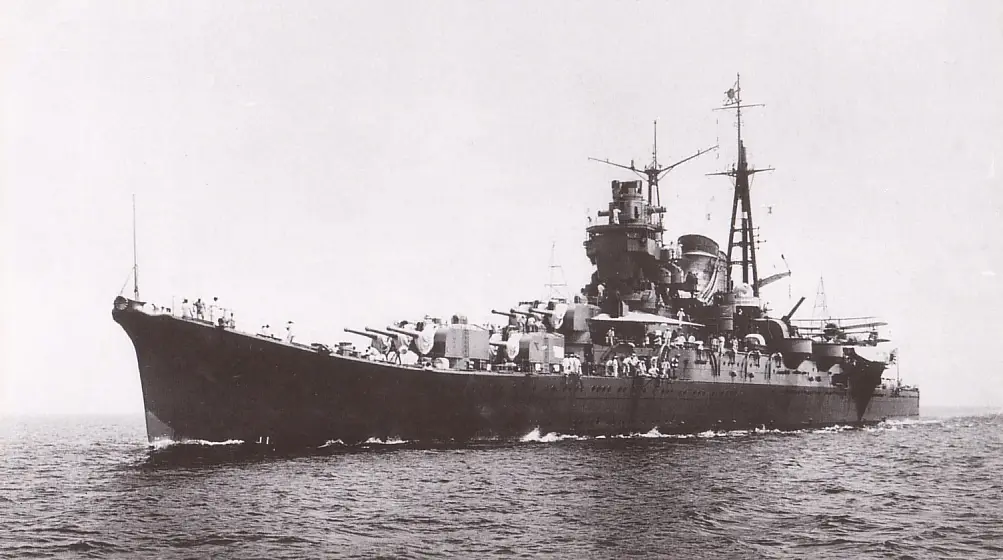 a black and white photograph shows a large, heavily armed battleship sailing on the ocean, with numerous crew members…