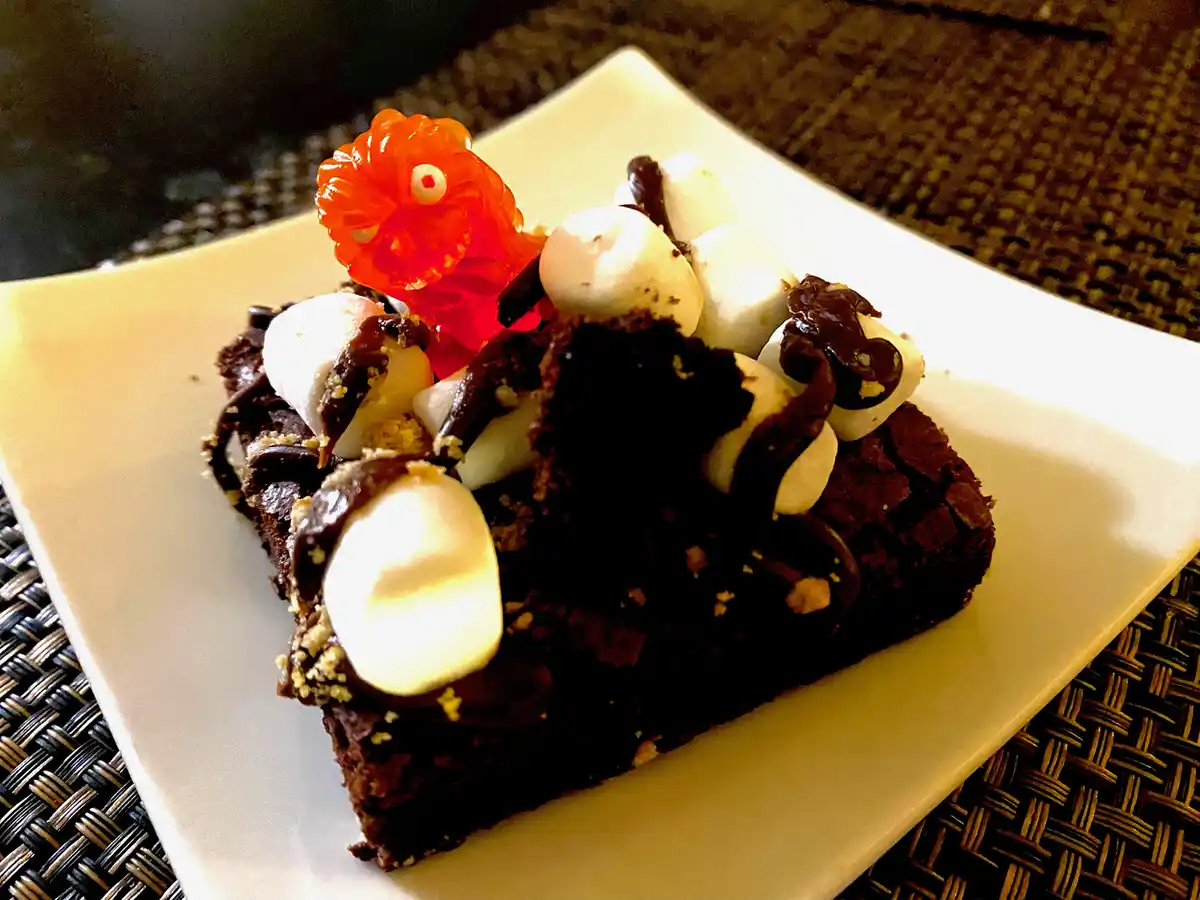 a close-up photo of a square chocolate brownie on a white plate, topped with melted chocolate, mini marshmallows, and a…