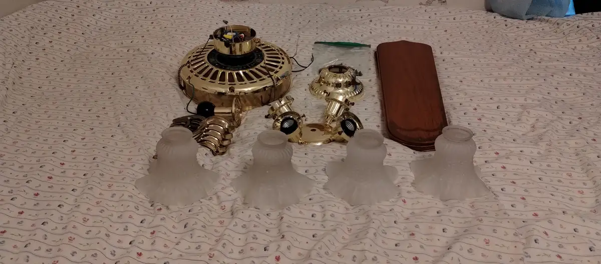 A disassembled ceiling fan with its gold motor housing, blades, light fixtures, and wooden cover laid out on a…