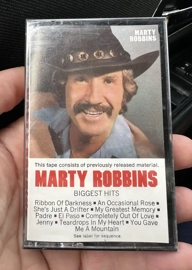 A hand holds a sealed cassette tape of Marty Robbins' 'Biggest Hits' album, featuring a photo of the singer in a cowboy hat…