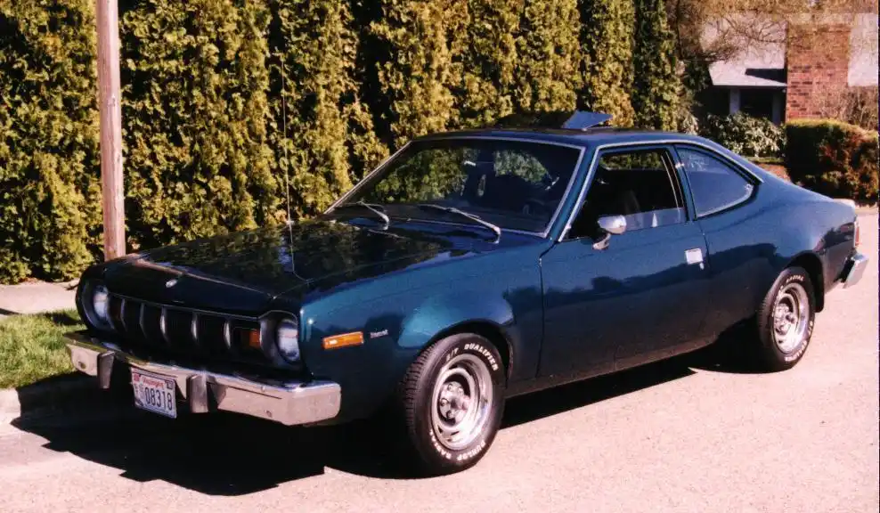A vintage teal AMC Pacer coupe is parked on a residential street with a hedge in the background. The car has a distinctive…