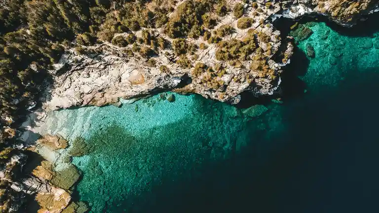 An aerial drone photo shows a stunning, clear turquoise cove bordered by rugged, tree-covered cliffs. The water transitions…
