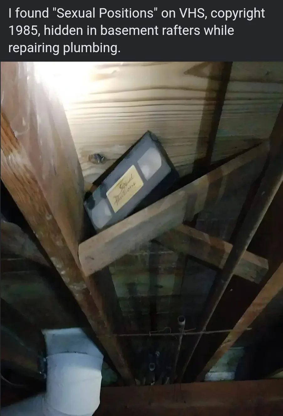 A VHS tape labeled "Sexual Positions" is wedged between wooden beams in a dimly lit basement crawlspace, with plumbing pipes…