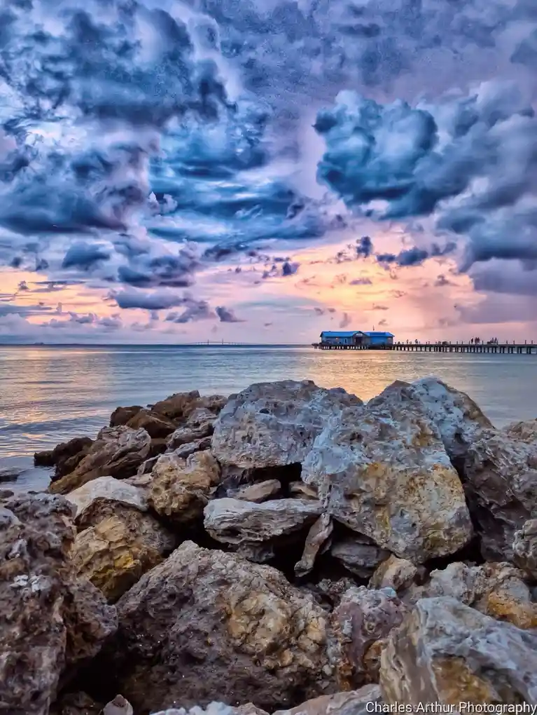 A scenic photograph shows a dramatic sky with swirling blue and purple clouds over a calm body of water at sunset, featuring…