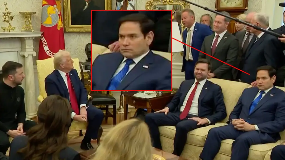 a still from a formal meeting shows a man in a blue suit and blue tie with a stern, unimpressed facial expression…