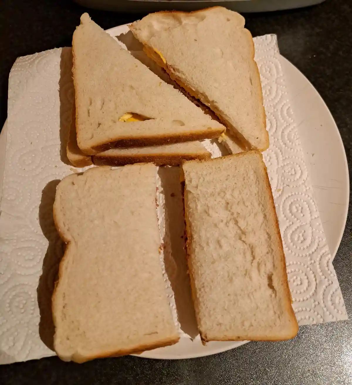 A white plate holds two sandwiches on a paper towel, one cut diagonally into two triangles and the other cut straight into…