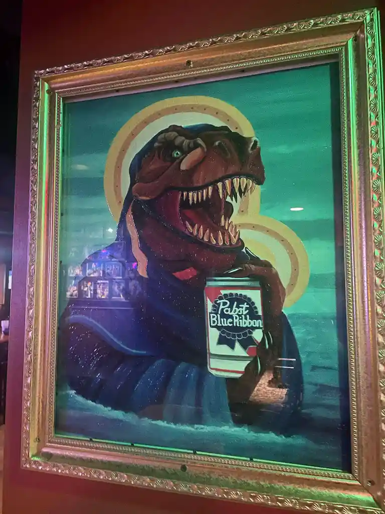 A framed painting depicts a T-Rex dressed as a saint, wearing a blue robe and holding a can of Pabst Blue Ribbon beer, with…