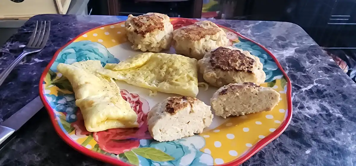 A colorful floral plate holds a folded omelet and five golden-brown biscuits, some whole and one halved, on a dark marble…