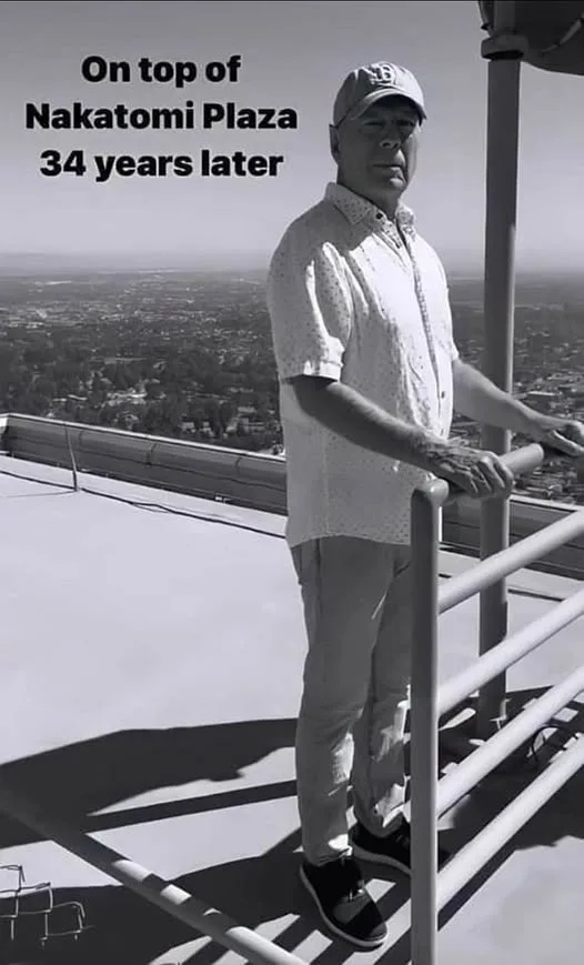 A black-and-white photo shows a man in a cap and light shirt standing on a rooftop with a cityscape behind him. He leans on…
