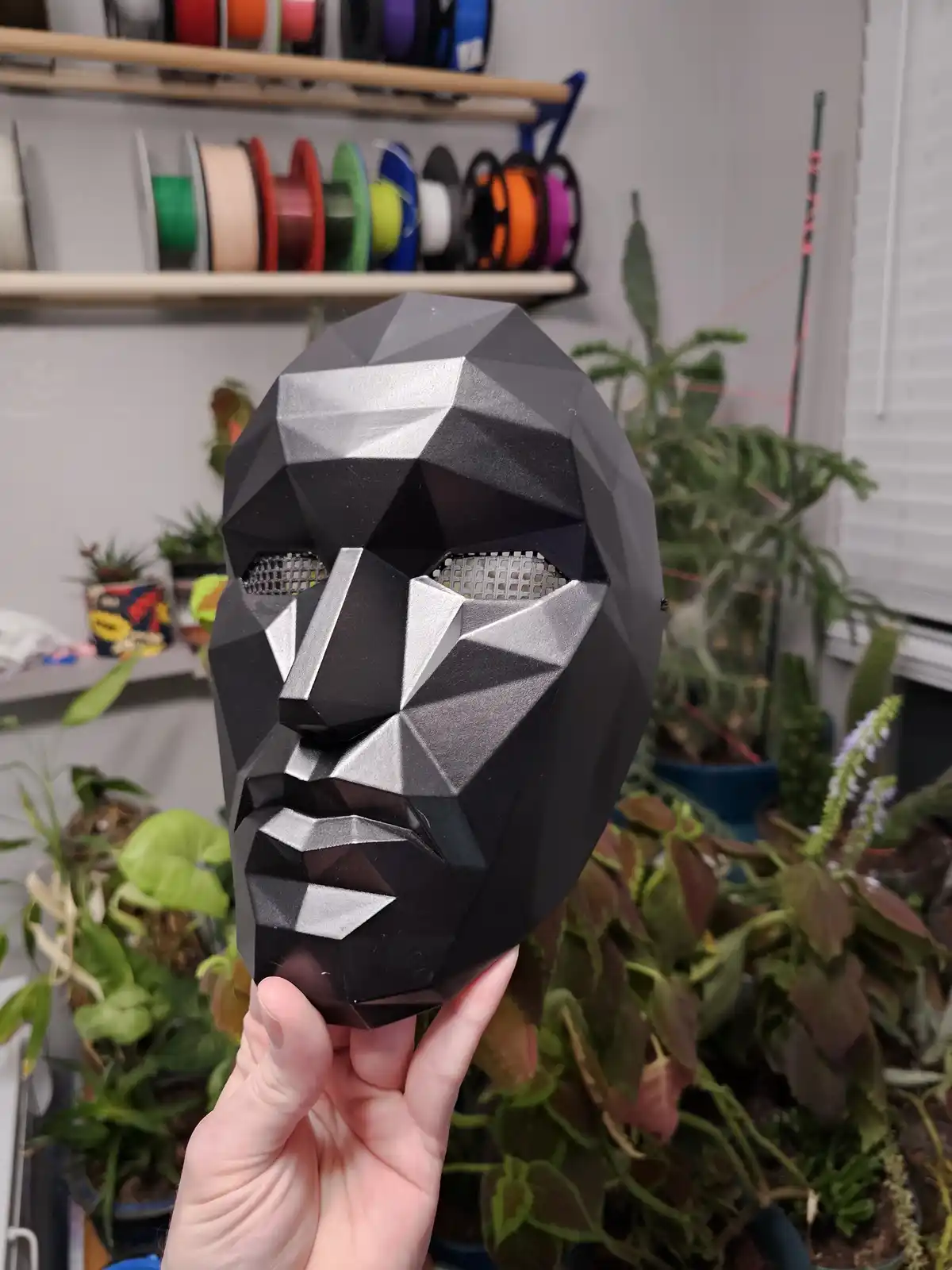 A hand holds up a black and silver geometrically faceted mask with mesh eye holes, set against a background of houseplants…