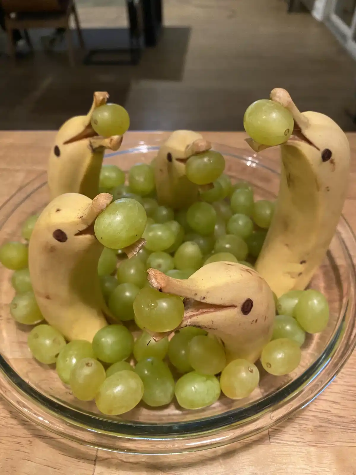 A clear glass bowl filled with green grapes and four bananas that have been creatively carved and arranged to resemble…