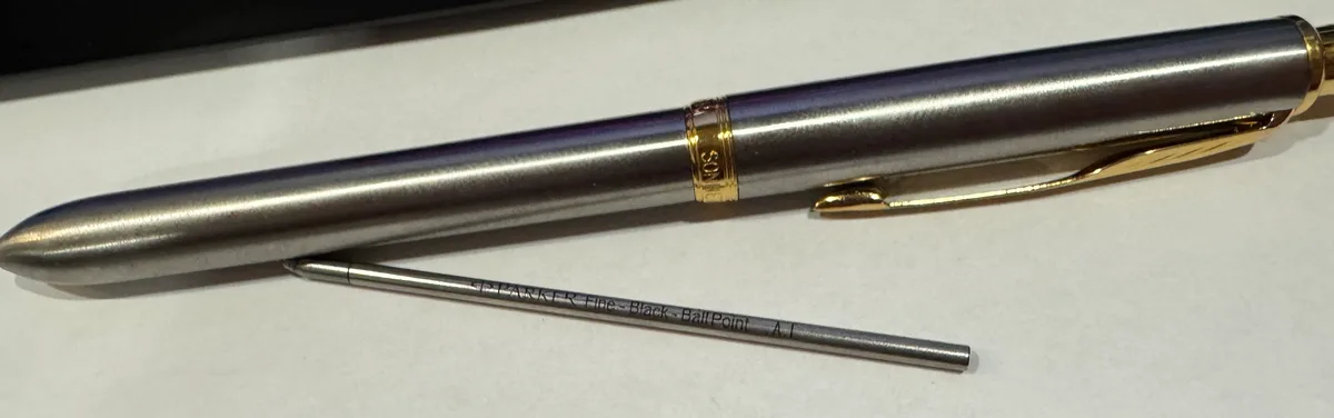 A close-up photo shows a sleek, silver Parker ballpoint pen with gold trim and clip, lying diagonally on a white surface…