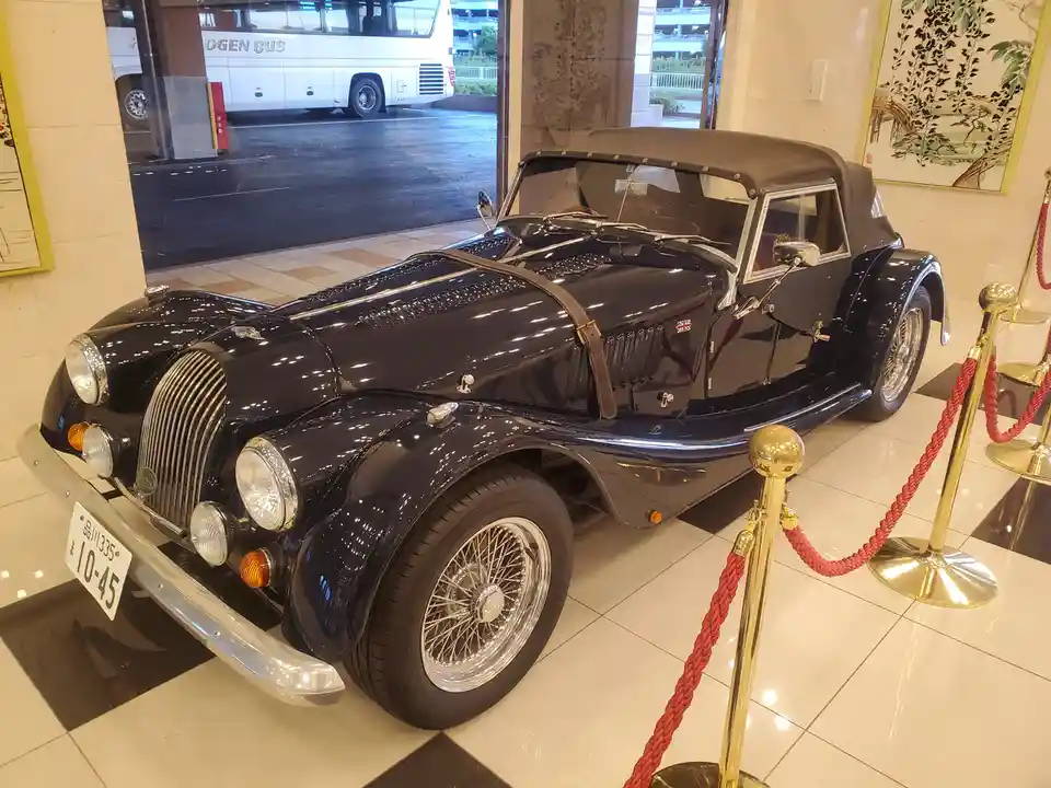 A glossy black vintage convertible car with wire wheels and a chrome grille is displayed indoors on a checkered floor, roped…