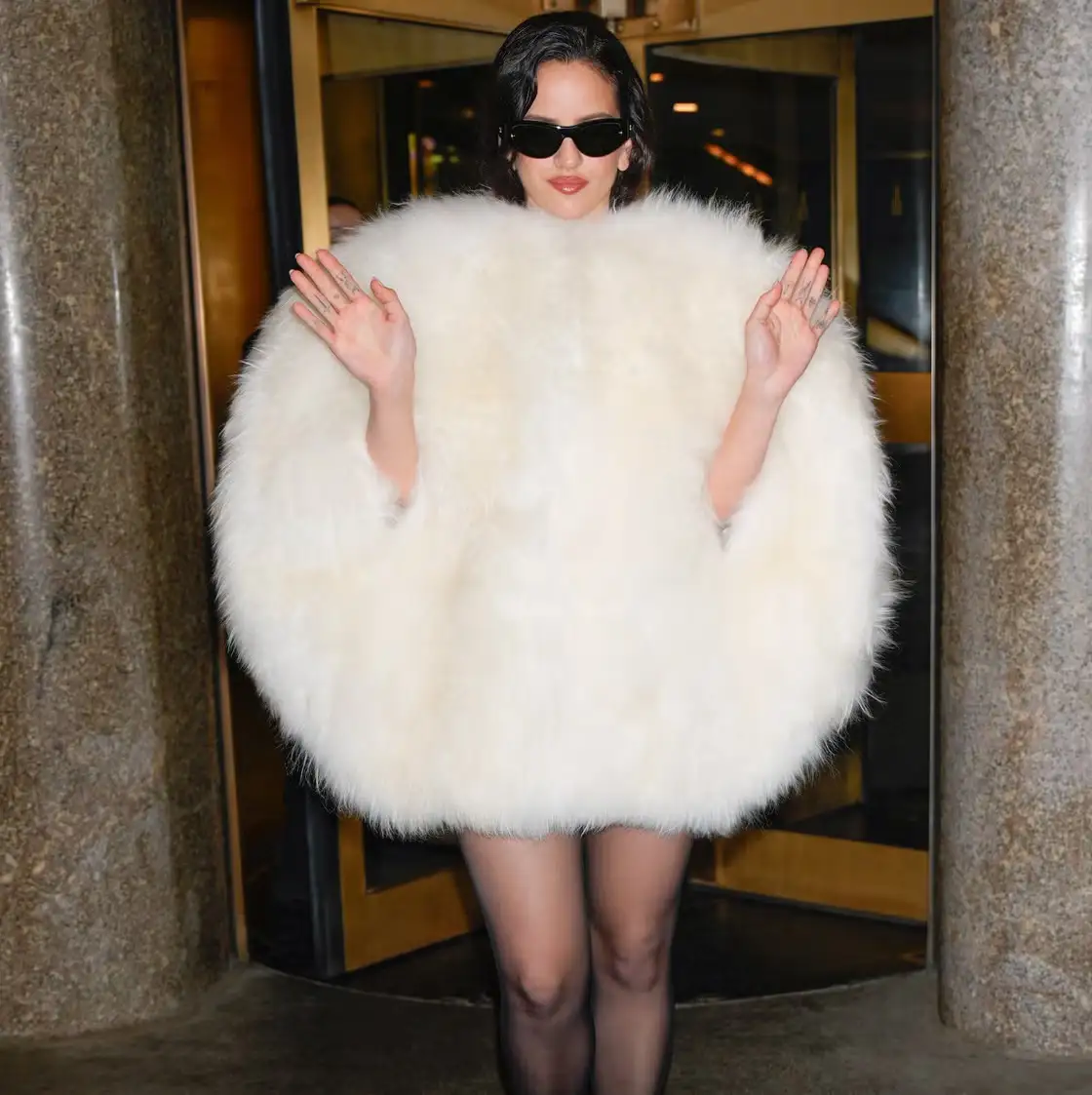 A woman stands in a doorway wearing a voluminous, fluffy white fur coat that obscures her torso and arms, with her hands…