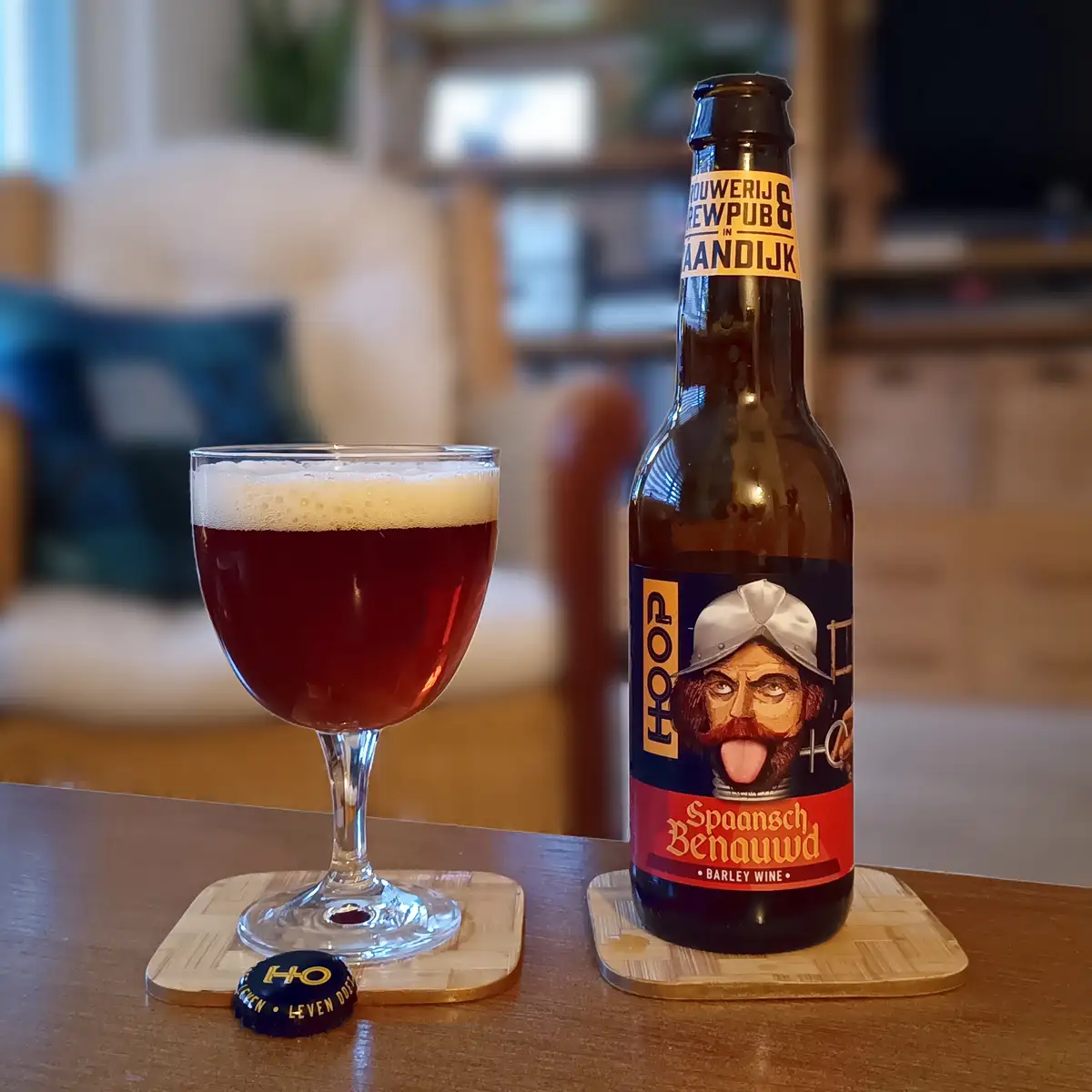A dark red beer in a stemmed glass with a foamy head sits next to its bottle, which features a label with a man in a helmet…