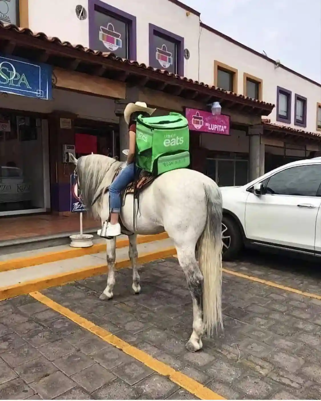A person wearing a cowboy hat and jeans is riding a white horse in a paved parking lot marked with yellow lines, carrying a…