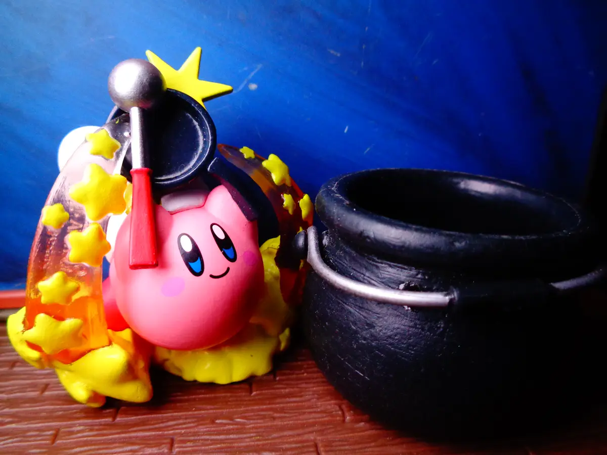 A pink Kirby figurine wearing a black witch's hat with a yellow star and a red tassel, surrounded by yellow stars, sits next…