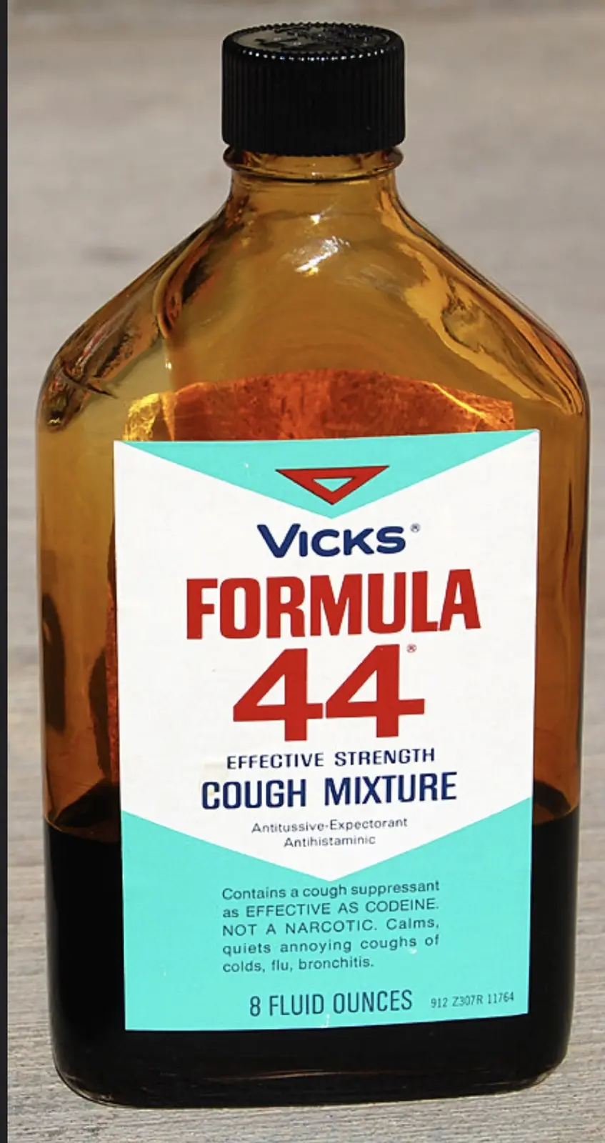 A close-up photo of an amber glass bottle of Vicks Formula 44 cough mixture with a black cap, sitting on a wooden surface…