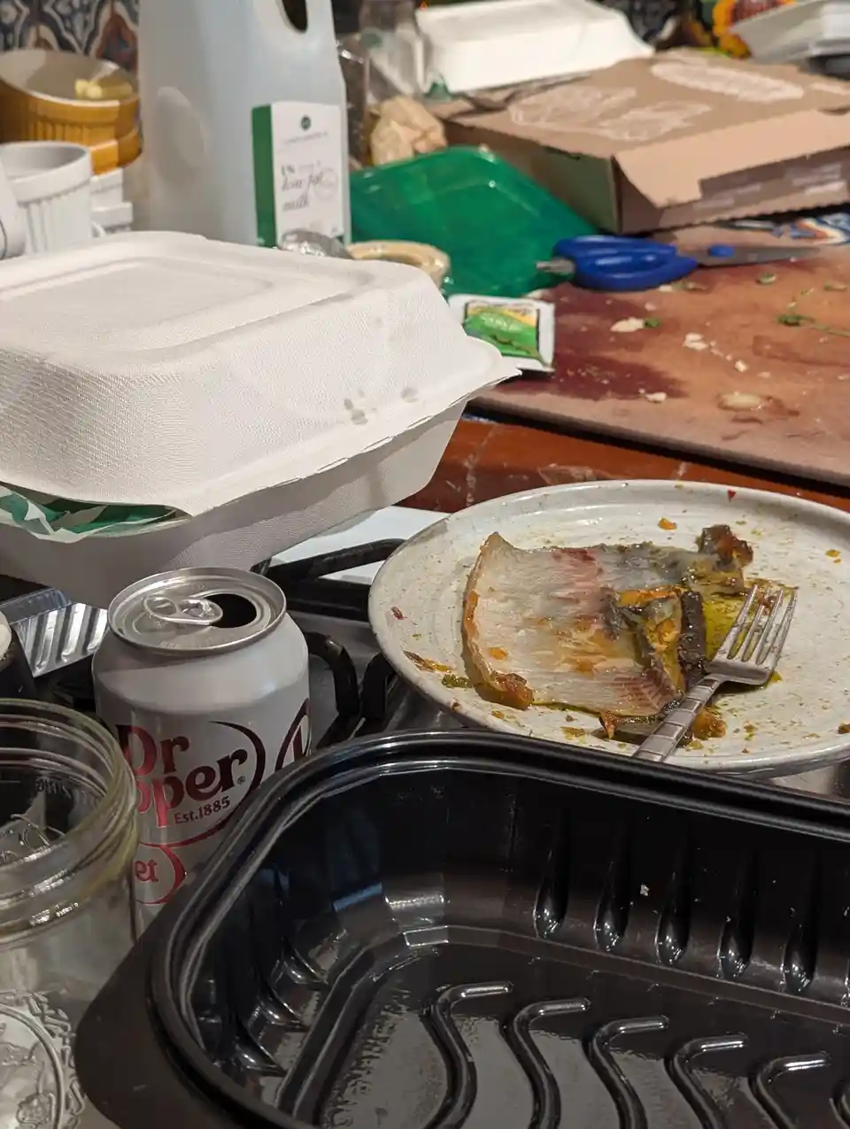 A messy kitchen counter shows a nearly empty plate with a single piece of cooked fish and a fork, next to a Dr Pepper can…