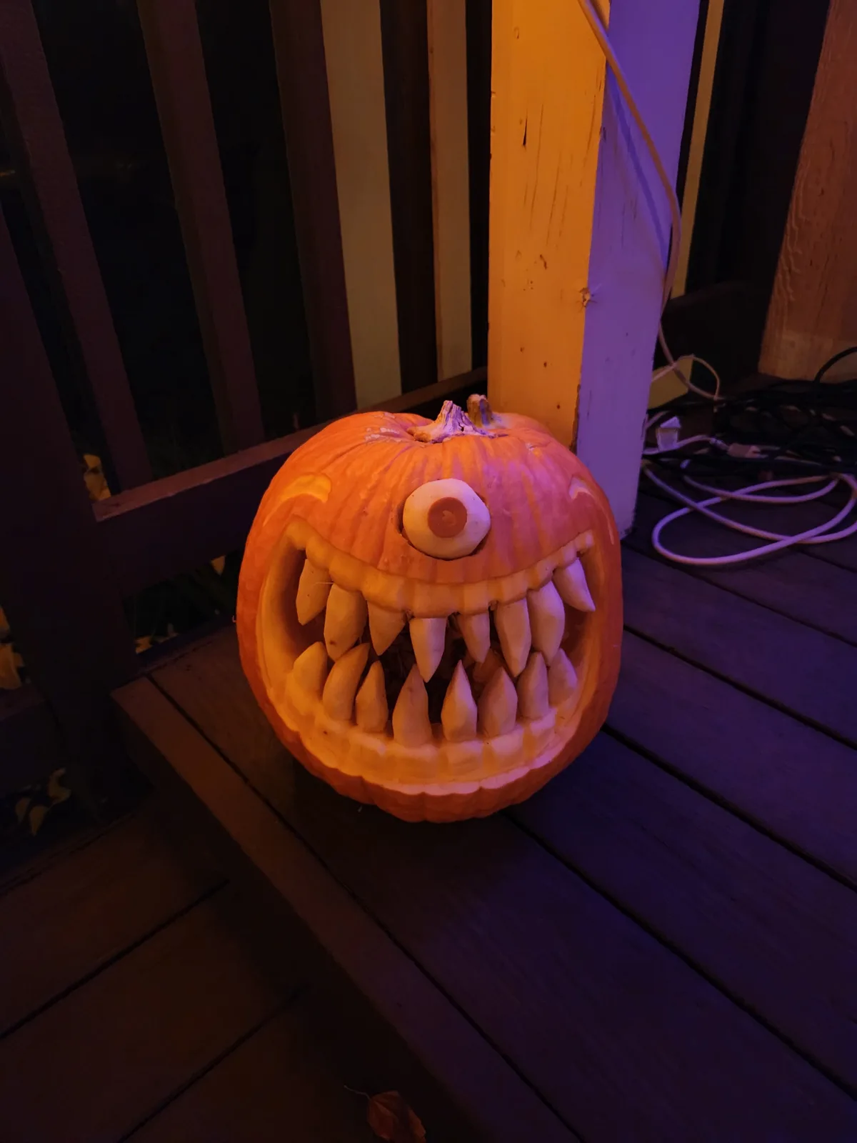 A carved Halloween pumpkin with a single large eye and sharp, jagged teeth sits on a wooden porch step, illuminated from…