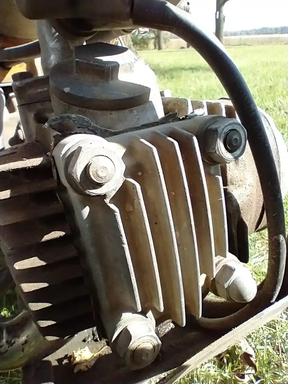 A close-up photo of a dusty, weathered engine block with visible bolts and fins, set outdoors on grass under bright…
