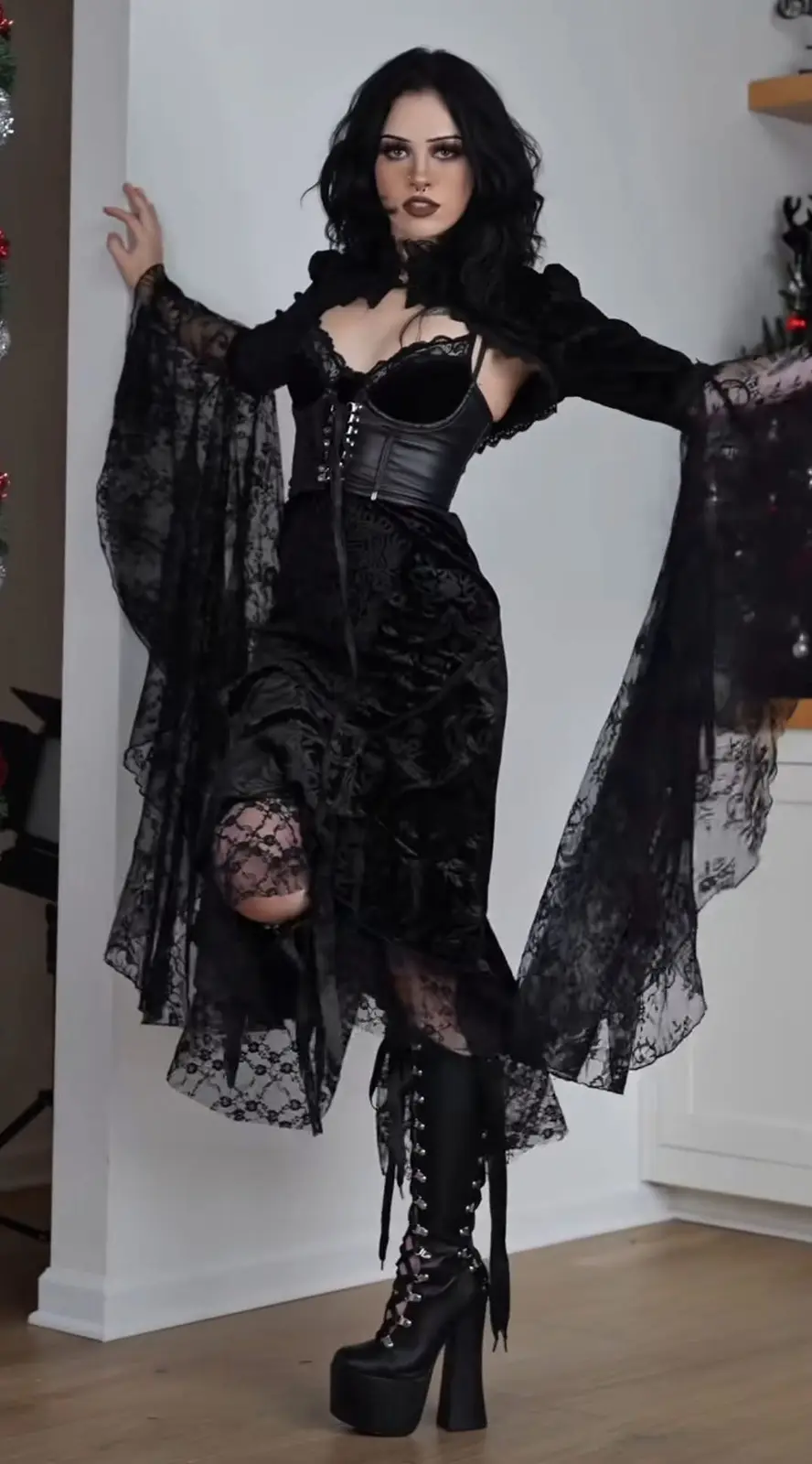 A woman with dark hair and gothic makeup poses indoors in a full black gothic outfit featuring lace, a corset, and platform…