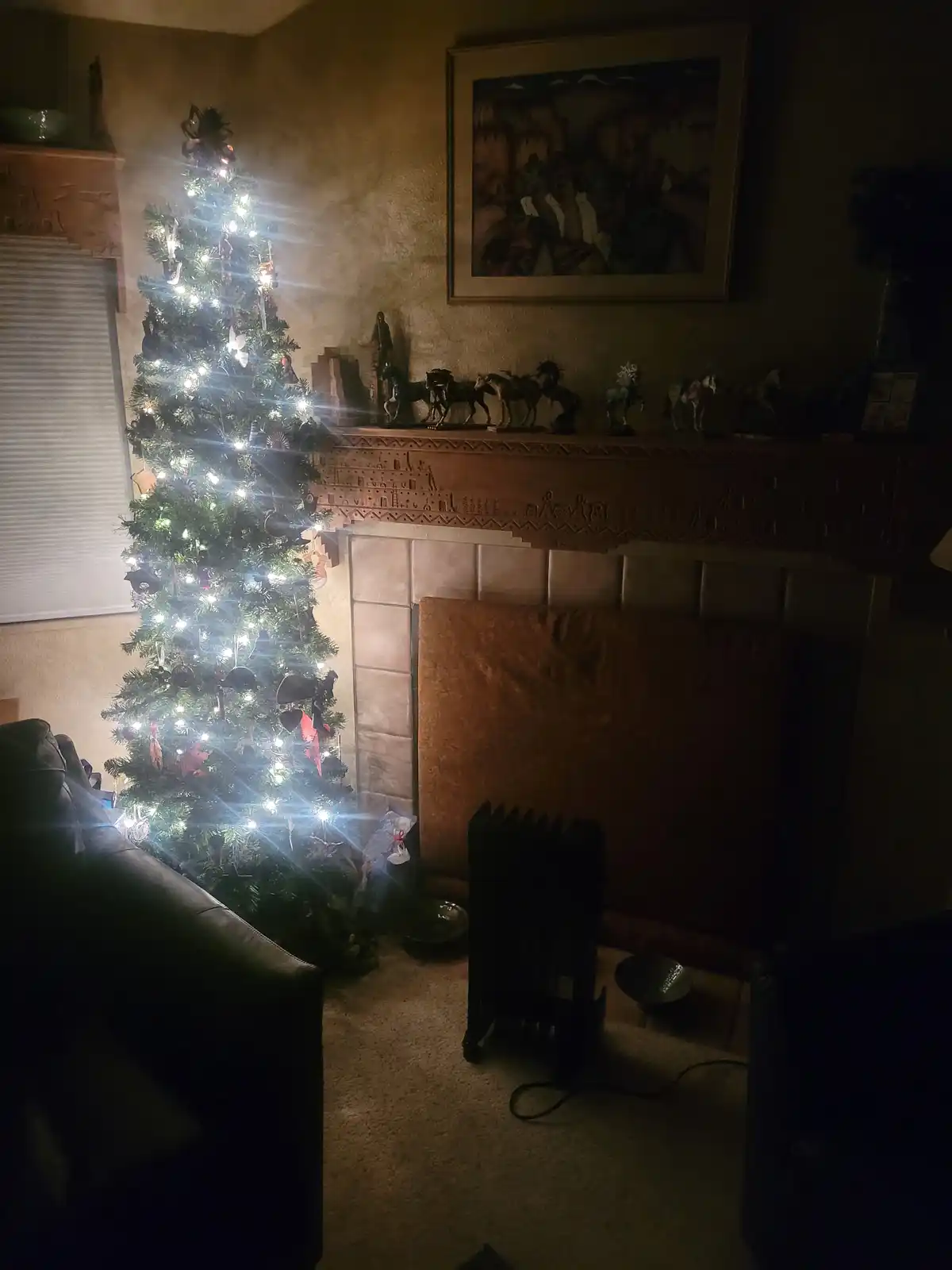 A dimly lit living room features a brightly lit, decorated Christmas tree standing directly in front of a fireplace mantel…