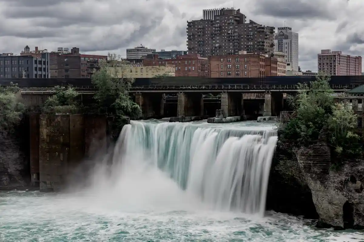 A long-exposure photograph of a wide, powerful waterfall cascading into a turbulent river, with a city skyline of brick and…