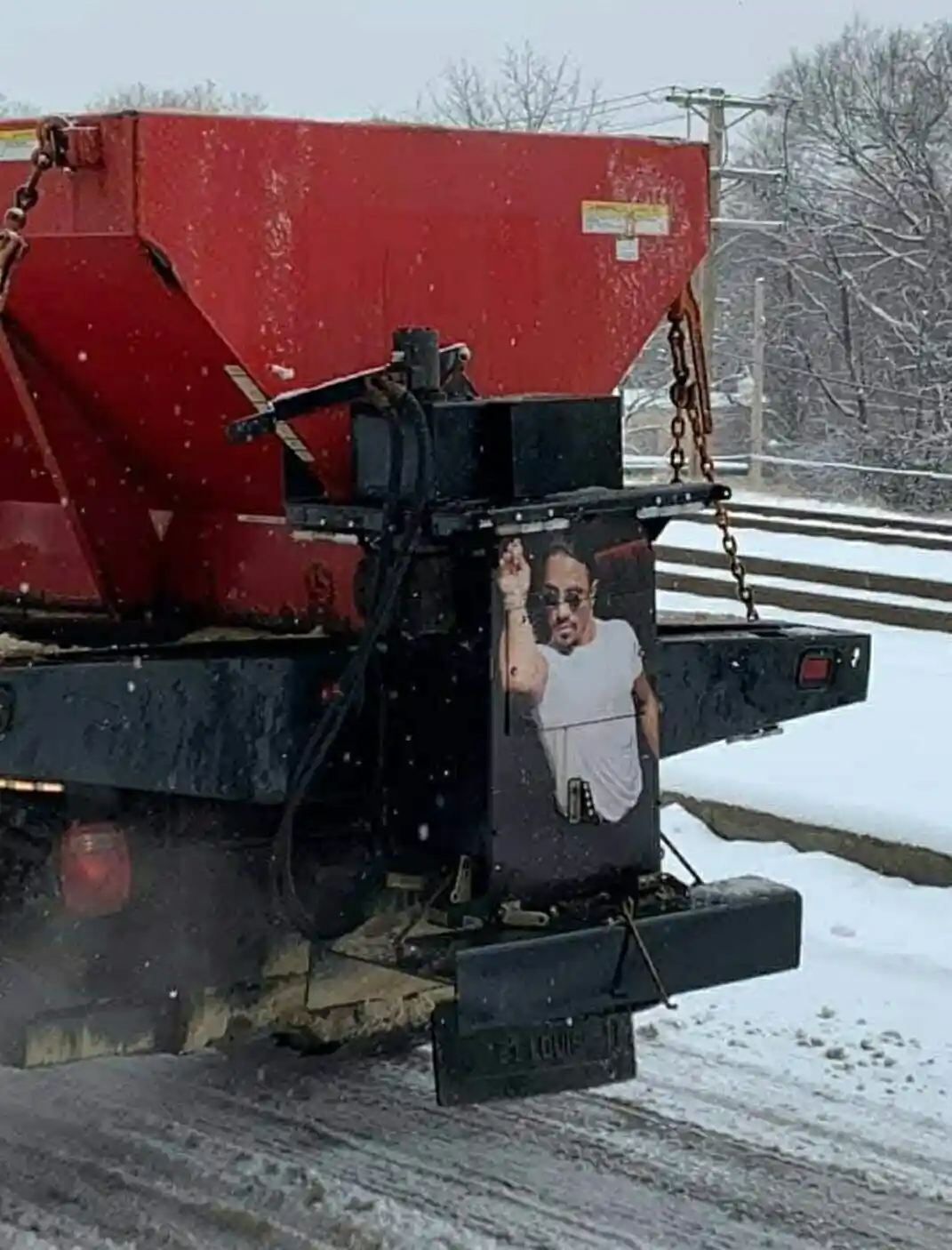 A red snowplow truck is shown in a snowy landscape, with a large printed image of the rapper 2Pac on its black front bumper…