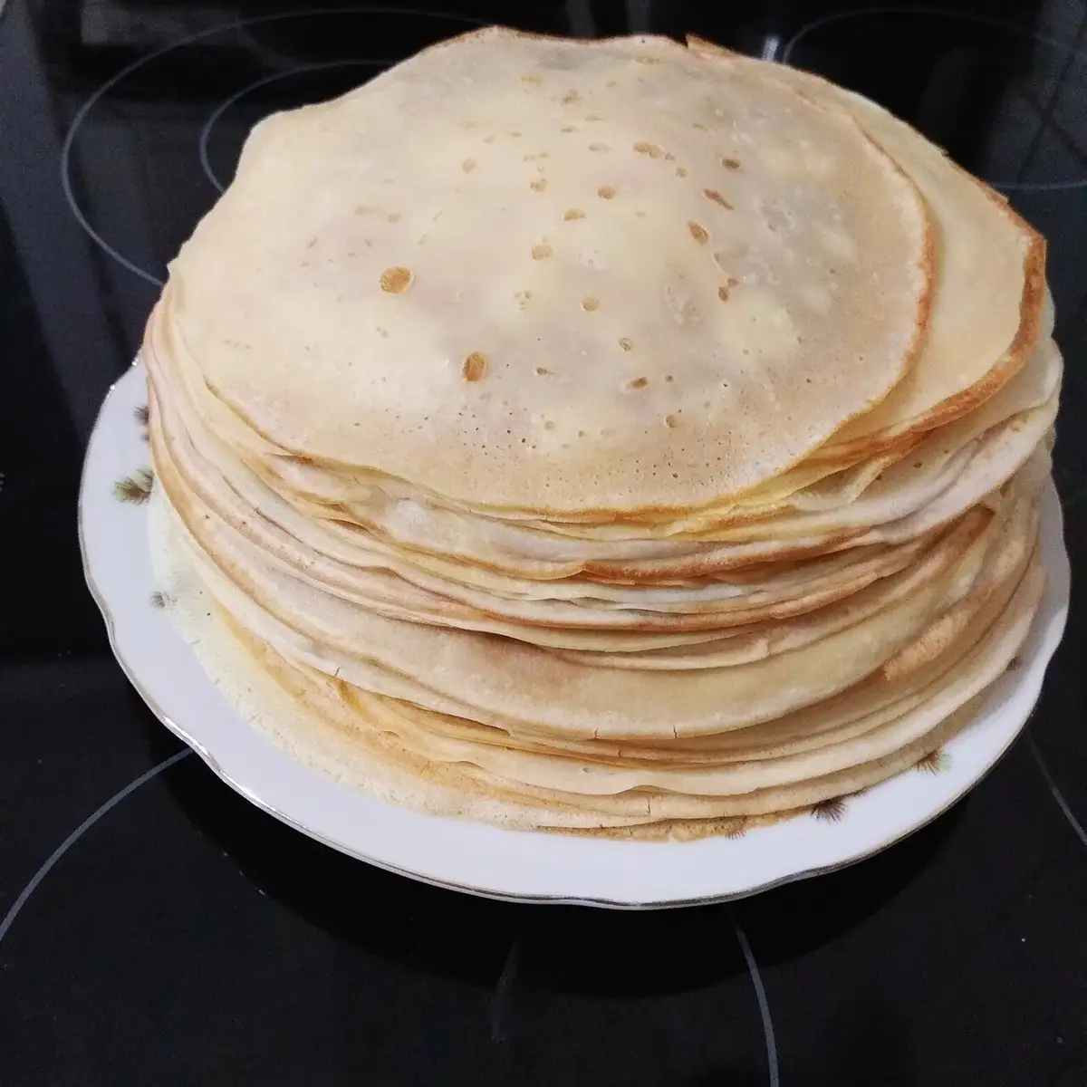 A tall, neat stack of thin, golden-brown crepes sits on a white plate with a decorative rim, placed on a black glass…