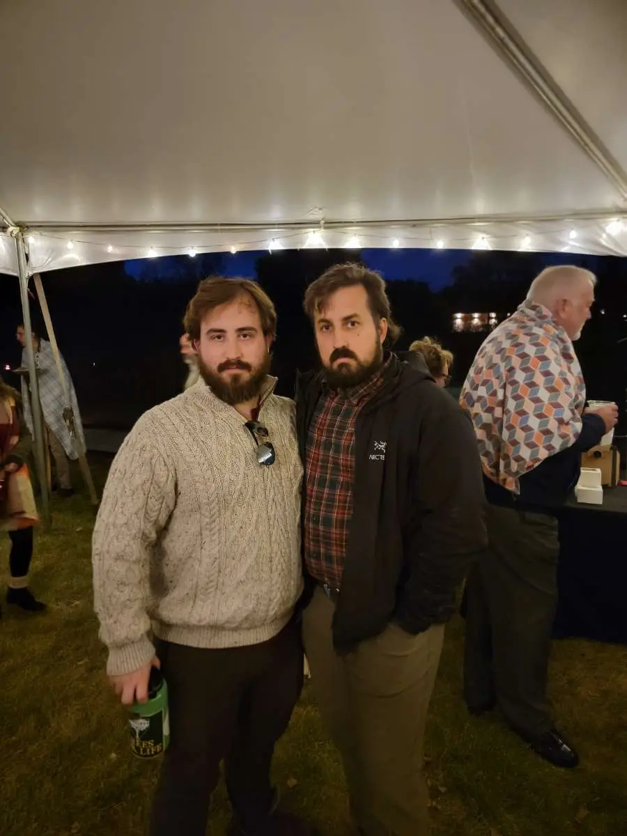 Two bearded men stand side-by-side under a tent at an outdoor evening event, with string lights above and other guests in…