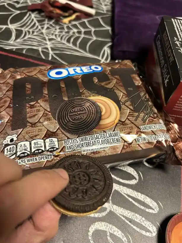 A hand holds an Oreo cookie in front of a package labeled 'OREO POST' with 'POSTY'S SWIRLED SALTED CARAMEL AND SHORTBREAD…