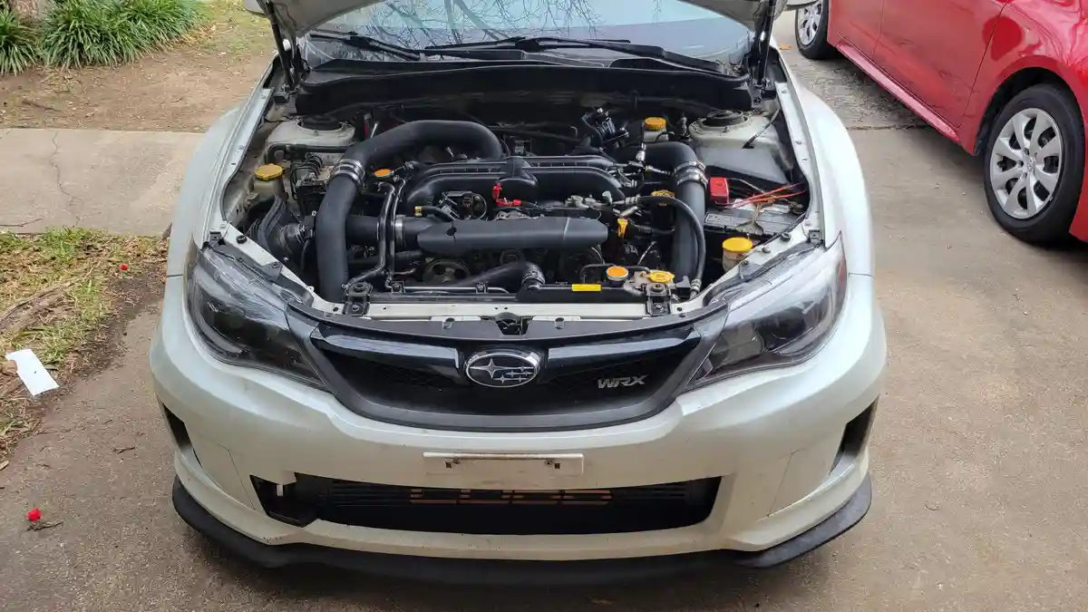 A front-facing photo of a white Subaru WRX with its hood open, revealing a modified engine bay with aftermarket parts. The…