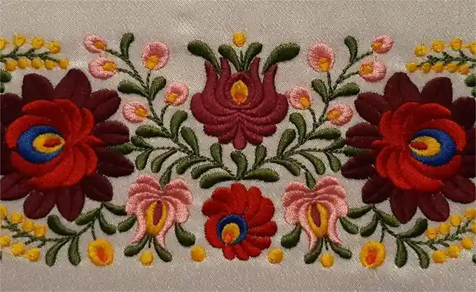A close-up of intricate floral embroidery on light-colored fabric, featuring large red and pink flowers with blue and yellow…