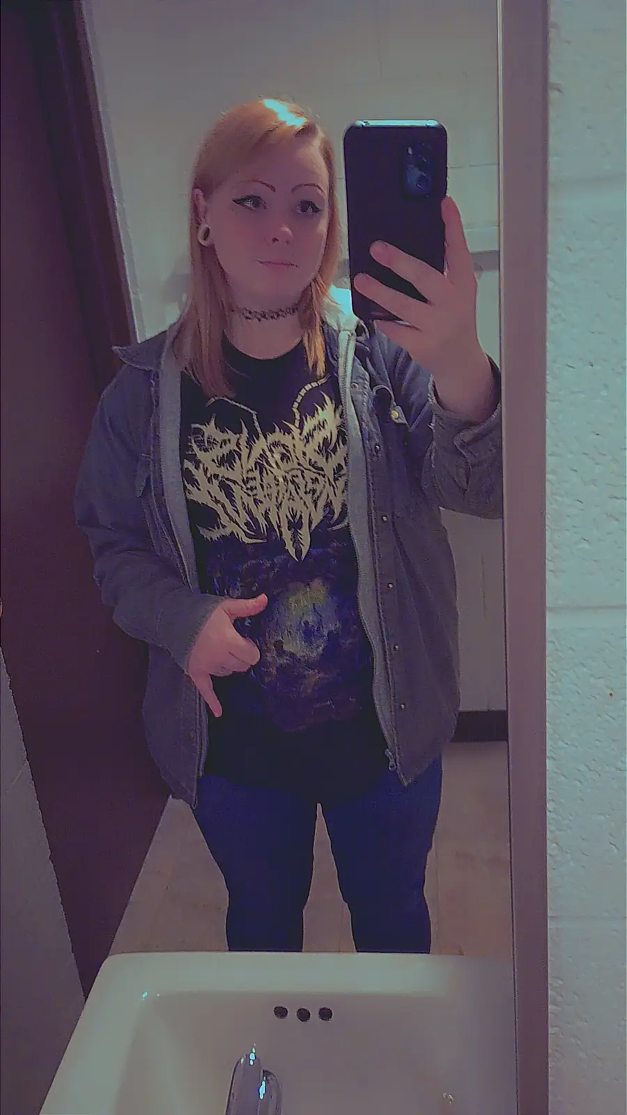 A person with light brown hair and a choker takes a mirror selfie in a bathroom, wearing a dark t-shirt with a white graphic…
