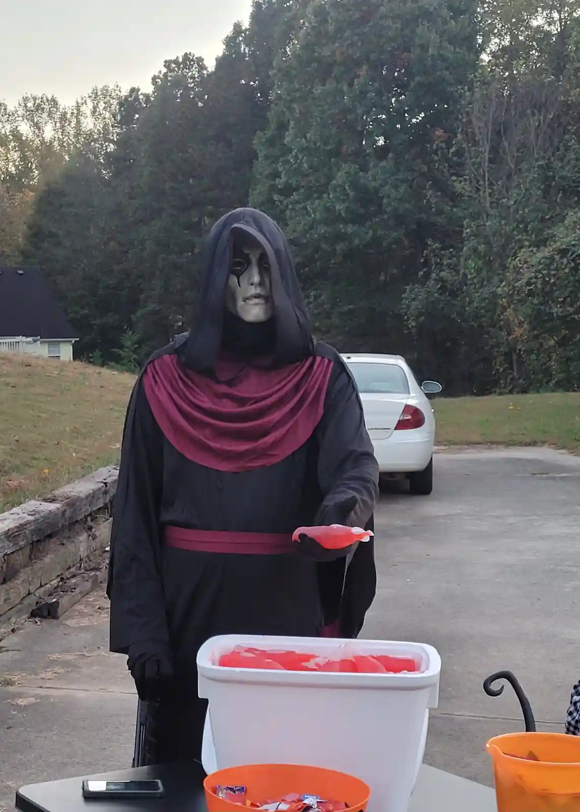 A person dressed as a grim reaper in a black robe with a red sash and hood stands behind a table outdoors, holding a small…