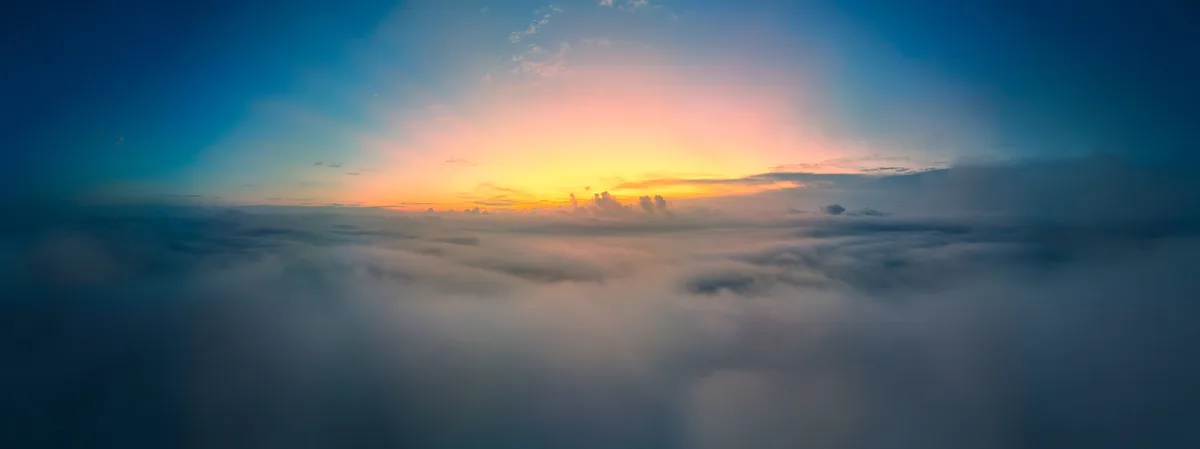 A panoramic view from above a sea of clouds during sunrise or sunset, showing a vibrant gradient sky transitioning from deep…