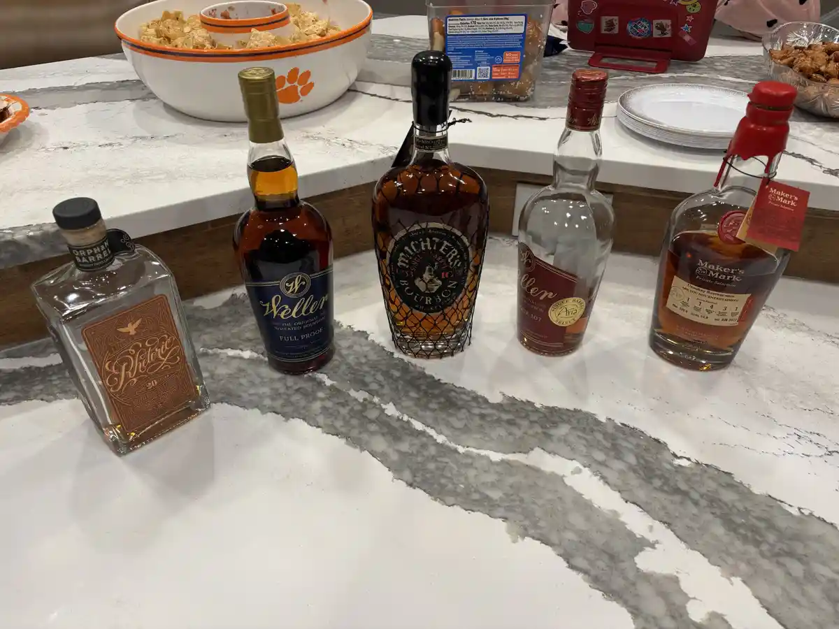 Five bottles of bourbon are lined up on a white marble countertop, with snacks and plates in the background. The bottles…