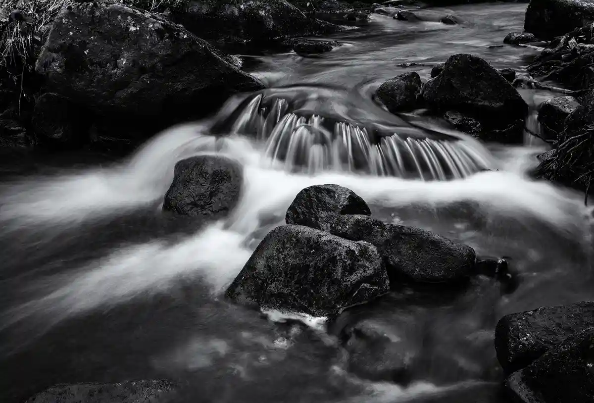 A black-and-white photograph of a flowing stream with smooth, blurred water cascading over dark, textured rocks. The image…